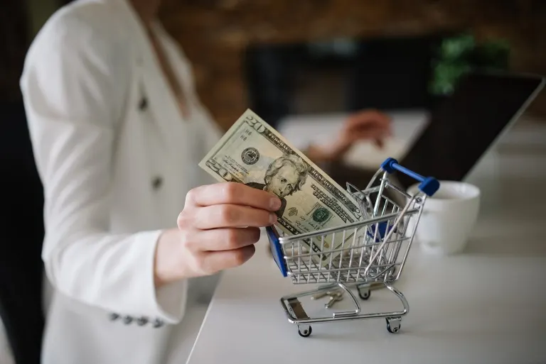Woman shops online with a small shopping cart and cash in a modern office setting during the afternoon